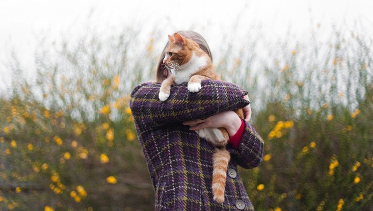 A blonde woman hugs her ginger cat outside. Her face is hidden by the cat.