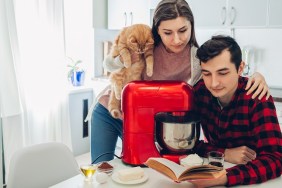 Young happy couple cooking on kitchen with food processor and holding cat. Woman and man reading recipe and hugging. Lifestyle