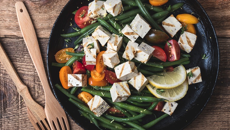 Delicious summer vegan meal, healthy green beans salad with grilled tofu, fresh colorful mix cherry tomatoes, thyme herbs and lemon zest served in rural cast iron skillet, wooden forks, top view