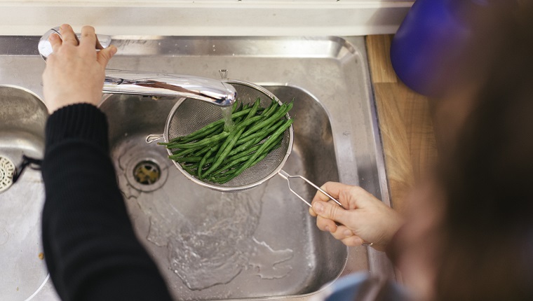 Close up of green beans being washed with water in a kitchen sink. They are being held under water by a woman that is not visible in the photo.