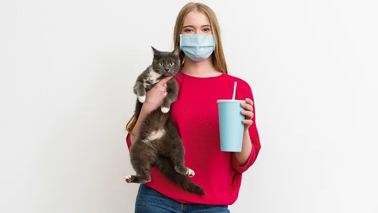 young woman in medical mask holding in arms cute cat and disposable cup with soda isolated on white