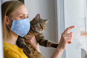 The woman put on a reusable protective mask for home isolation from coronavirus and covid-19. A girl holds a Cat in her arms, indoors or at home by the window. Preventing the spread of the virus and disease. Waiting for the quarantine to complete.