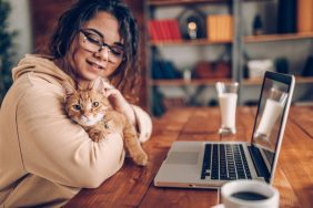 Woman in a hoodie cuddling with orange cat while she is working from home on her laptop at the kitchen table.