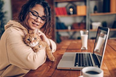 Woman in a hoodie cuddling with orange cat while she is working from home on her laptop at the kitchen table.