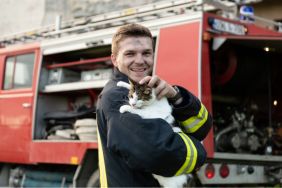 Close-up portrait of heroic fireman in protective suit and red helmet holds saved cat in his arms.