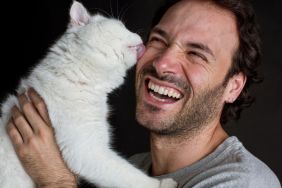 White cat licks man's face as he laughs happily.